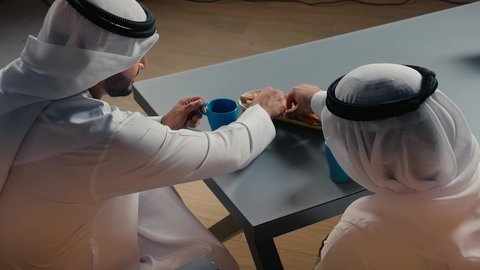 The concept of entertainment and leisure, spending a good time with friends, having a quick meal and a refreshing drink, a close-up shot from the back of two Emirati Gulf Arab men wearing kandura and ghutra exchanging conversations and discussions inside a cafe with gestures of pleasure and happiness