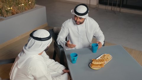 Having a good time with friends, having a quick meal and a refreshing drink, two Emirati Gulf Arab men wearing kandura and ghutra exchanging conversations and discussions inside a cafe with gestures of pleasure and happiness, the concept of entertainment and recreation
