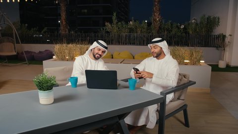 النقاش والحوار بين زملاء العمل، رجلان عربيان خليجيان اماراتيان يرتديان الكندورة و الغنتره في اجتماع عمل خارج الشركة يقومان بالعمل على الحاسب المحمول، مفهوم التكنولوجيا وإستخدام أجهزة تقنية حديثة، ايماءات الاندماج و التركيز ، قضاء وقت ممتع مع الأصدقاء في عطلة نهاية الأسبوع ، الاستمتاع بتناول المشروبات
