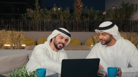 زملاء عمل عرب خليجيون اماراتيون يرتدون الكندورة و الغنتره في اجتماع عمل خارج الشركة يقومان بالعمل على الحاسب المحمول، مفهوم التكنولوجيا وإستخدام أجهزة تقنية حديثة، ايماءات الاندماج و التركيز ، قضاء وقت ممتع مع الأصدقاء في عطلة نهاية الأسبوع ، الاستمتاع بتناول المشروبات