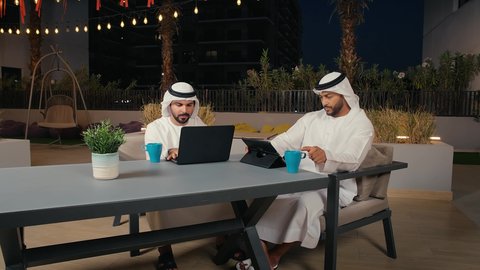 مفهوم التكنولوجيا وإستخدام أجهزة تقنية حديثة، ايماءات الاندماج و التركيز ، قضاء وقت ممتع مع الأصدقاء في عطلة نهاية الأسبوع,, زملاء عمل عرب خليجيون اماراتيون في اجتماع عمل خارج الشركة يقومان بالعمل على التابلت و الحاسب المحمول ، الاستمتاع بتناول المشروبات