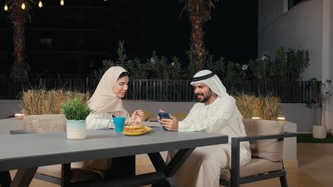 متابعة مواقع التواصل الاجتماعي عبر الهاتف المحمول، تصفح تطبيقات الموبايل، رجل عربي خليجي اماراتي يرتدي الكندورة والغترة يقوم بتصفح الجوال برفقه زوجته داخل احد المقاهي الاماراتية، دمج التقنيات الحديثة بالحياة العملية