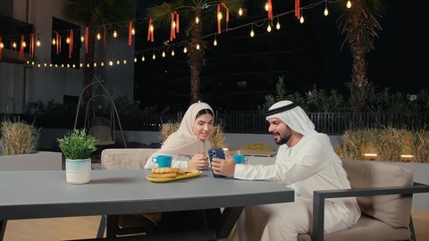 Browsing mobile applications, an Emirati Gulf Arab man wearing a kandura and ghutra browsing his mobile phone with his wife inside an Emirati cafe, following social media sites via mobile phone, integrating modern technology into practical life.