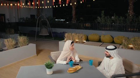 قضاء وقت ممتع في الخارج، زوجان عربيان خليجيان اماراتيان يرتديان الزي التقليدي يستمتعان بتبادل الحديث في احد المقاهي الاماراتية، نقاشات وحوارات، أجواء عائلية سعيدة، الاستمتاع بتناول المشروبات