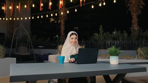 Completing work tasks remotely via the internet, making video calls via laptop, a young Arab Gulf Emirati woman working on the laptop with gestures of happiness and pleasure, using modern and advanced technology, a Saudi woman sitting in a cafe in the United Arab Emirates