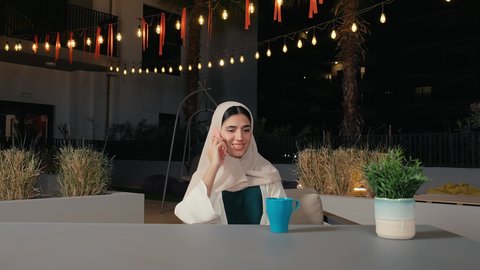 Making a phone call using a mobile phone, chatting with friends, a veiled Emirati Gulf Arab woman wearing an abaya sitting in a restaurant with gestures of happiness and pleasure, a Saudi woman enjoying a drink and having a good time outside
