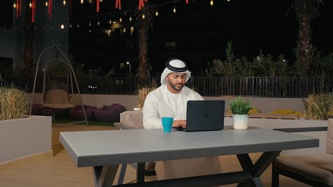 Accomplishing tasks in a calm and comfortable atmosphere, enjoying drinks, using modern technology and techniques, following up on work remotely via laptop, an Arab Gulf Emirati man wearing a kandura and ghutra sitting outdoors in a restaurant in the United Arab Emirates