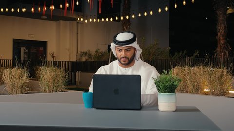 الاستمتاع بتناول المشروبات, إنجاز المهام في جو هادئ ومريح، استخدام التكنولوجيا والتقنيات الحديثة، متابعة العمل عن بعد عبر الحاسوب المحمول، رجل عربي خليجي اماراتي يرتدي الكندورة و الغتره يجلس في الهواء الطلق في أحد المطاعم بالامارات العربية