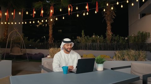 اجتماع عمل عن بعد، استخدم جهاز الحاسوب، تصوير مقرب من الجانب لرجل عربي خليجي إماراتي يجلس فى احد المقاهي في الهواء الطلق يرفع يديه بايماءات القاء التحية والسلام ، إجراء مكالمات فيديو، تواصل اجتماعي، محادثات ودردشة، التواصل مع الأصدقاء، تصفح الانترنت