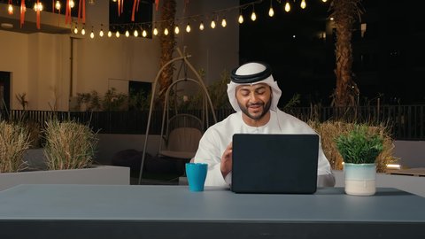 استخدم جهاز الحاسوب، رجل عربي خليجي إماراتي يجلس فى احد المقاهي في الهواء الطلق يرفع يديه بايماءات القاء التحية والسلام ، إجراء مكالمات فيديو، تواصل اجتماعي، محادثات ودردشة، التواصل مع الأصدقاء، اجتماع عمل عن بعد، تصفح الانترنت