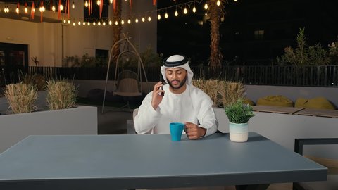 التواصل مع الأهل والأصدقاء، استخدام التقنيات والأجهزة الحديثة، إيماءات وتعابير السعادة والسرور لسماع الأخبار المفرحة، تصوير مقرب لرجل عربي خليجي إماراتي مبتسم يرتدي الكندورة والغترة يقوم يجلس في المقهي ويقوم بإجراء مكالمة هاتفية عبر الهاتف المحمول، الاستمتاع بتناول المشروبات