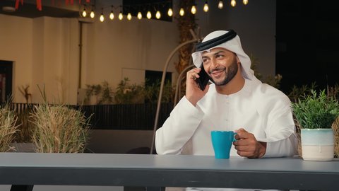 استخدام التقنيات والأجهزة الحديثة، التواصل مع الأهل والأصدقاء، إيماءات وتعابير السعادة والسرور لسماع الأخبار المفرحة، تصوير مقرب لرجل عربي خليجي إماراتي مبتسم يرتدي الكندورة والغترة يقوم يجلس في المقهي ويقوم بإجراء مكالمة هاتفية عبر الهاتف المحمول، الاستمتاع بتناول المشروبات