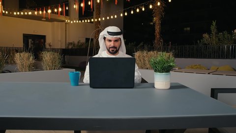 إنجاز المهام في جو هادئ ومريح، استخدام التكنولوجيا والتقنيات الحديثة، متابعة العمل عن بعد عبر الحاسوب المحمول، رجل عربي خليجي اماراتي يرتدي الكندورة و الغتره يجلس في الهواء الطلق في أحد المطاعم بالامارات العربية، الاستمتاع بتناول المشروبات