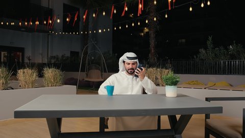 إجراء مكالمة هاتفية باستخدام الموبايل، التسامر والدردشة مع الأصدقاء، رجل عربي خليجي اماراتي يرتدي الكندورة و الغتره يجلس في المطعم بايماءات السعادة و السرور، الاستمتاع  بتناول المشروبات و بقضاء وقت ممتع فى الخارج