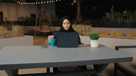 استخدام التكنولوجيا والتقنيات الحديثة، متابعة العمل عن بعد عبر الحاسوب المحمول، امرأة عربية خليجية اماراتية ترتدي العباءة والحجاب تجلس في الهواء الطلق في أحد المطاعم بالامارات العربية،سيده سعودية تقوم بإنجاز المهام في جو هادئ ومريح، الاستمتاع بتناول المشروبات