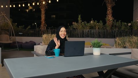 التواصل مع العائلة و الأصدقاء في أوقات الفراغ، إجراء مكالمة فيديو عبر الحاسب المحمول، مشاركة الأخبار والأحاديث، امراه عربيه خليجيه اماراتية ترتدي العباءة  تستمع بقضاء وقت ممتع فى المطعم، سيده سعودية تقوم بإلقاء التحية والسلام، تعابير الترحيب والاستقبال