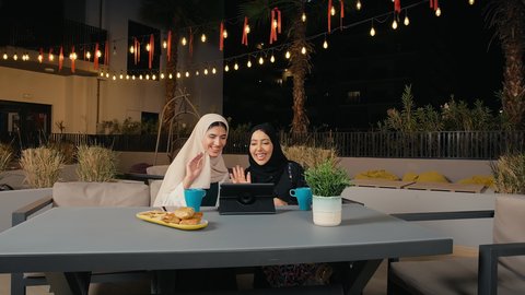 تطوير العلاقات الإجتماعية، إجراء مكالمة فيديو باستخدام التابلت، جلسة وناسة يوم الاجازة، كوفي شوب راقي بالامارات، التسامر والدردشة مع الأصدقاء، تعابير السعادة والسرور بلمة الرفاق، امرأة عربية خليجية اماراتية محجبة تجلس مع صديقتها في المطعم، صديقتان سعوديتان يستمتعان بقضاء وقت ممتع فى الخارج