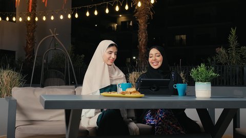 إجراء مكالمة فيديو باستخدام التابلت، جلسة وناسة يوم الاجازة، كوفي شوب راقي بالامارات، تطوير العلاقات الإجتماعية، التسامر والدردشة مع الأصدقاء، تعابير السعادة والسرور بلمة الرفاق، امرأة عربية خليجية اماراتية محجبة تجلس مع صديقتها في المطعم، صديقتان سعوديتان يستمتعان بقضاء وقت ممتع فى الخارج