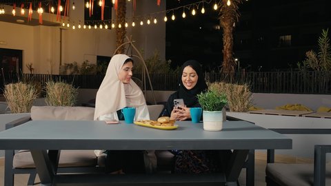 استخدام الاجهزة التقنية الحديثة، جلسة وناسة يوم الاجازة، كوفي شوب راقي بالامارات، تطوير العلاقات الإجتماعية، التسامر والدردشة مع الأصدقاء، تعابير السعادة والسرور بلمة الرفاق، امرأة عربية خليجية اماراتية محجبة تجلس مع صديقتها في المطعم تمسك بيدها الهاتف المحمول، صديقتان سعوديتان يستمتعان بقضاء وقت ممتع فى الخارج