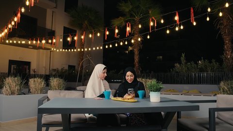 إجراء مكالمة هاتفية باستخدام الموبايل، جلسة وناسة يوم الاجازة، كوفي شوب راقي بالامارات، تطوير العلاقات الإجتماعية، التسامر والدردشة مع الأصدقاء، تعابير السعادة والسرور بلمة الرفاق، امرأة عربية خليجية اماراتية محجبة تجلس مع صديقتها في المطعم، صديقتان سعوديتان يستمتعان بقضاء وقت ممتع فى الخارج