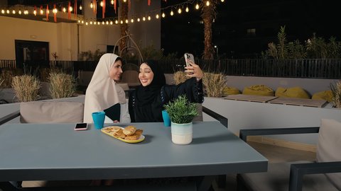 جلسة وناسة يوم الاجازة، التقاط صور السيلفي برفقة الأصدقاء من خلال الهاتف المحمول، كوفي شوب راقي بالامارات، تطوير العلاقات الإجتماعية، التسامر والدردشة مع الأصدقاء، تعابير السعادة والسرور بلمة الرفاق، امرأة عربية خليجية اماراتية محجبة تجلس مع صديقتها في المطعم، صديقتان سعوديتان يستمتعان بقضاء وقت ممتع فى الخارج