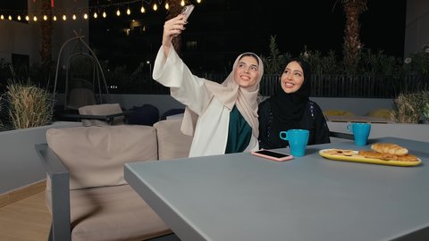 التقاط صور السيلفي برفقة الأصدقاء من خلال الهاتف المحمول، جلسة وناسة يوم الاجازة، كوفي شوب راقي بالامارات، تطوير العلاقات الإجتماعية، التسامر والدردشة مع الأصدقاء، تعابير السعادة والسرور بلمة الرفاق، امرأة عربية خليجية اماراتية محجبة تجلس مع صديقتها في المطعم، صديقتان سعوديتان يستمتعان بقضاء وقت ممتع فى الخارج