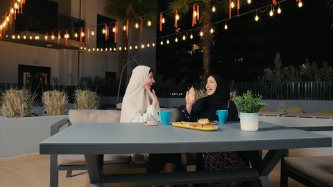 الاستمتاع بالمشروبات الباردة، جلسة وناسة يوم الاجازة، كوفي شوب راقي بالامارات، تطوير العلاقات الإجتماعية، التسامر والدردشة مع الأصدقاء، تعابير السعادة والسرور بلمة الرفاق، امرأة عربية خليجية اماراتية محجبة تجلس مع صديقتها في المطعم، صديقتان سعوديتان تتبادلان الحديث