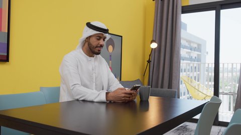 الدردشة عبر الانترنت ،استخدام التقنيات والتكنولوجيا الحديثة ، قضاء وقت ممتع ، الاطمئنان على الاهل والاصدقاء ، الاستمتاع بالمشروبات الساخنه ، تصوير لشاب عربي خليجي إماراتي يرتدي الكندورة والغترة البيضاء يجلس على اريكة رمادية ، يستخدم جهاز الهاتف المحمول