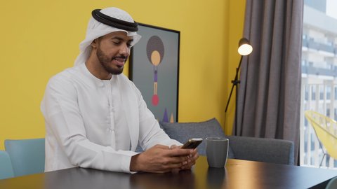 قضاء وقت ممتع ، الاطمئنان على الاهل والاصدقاء ، الاستمتاع بالمشروبات الساخنه ،  الدردشة عبر الانترنت ،دمج التكنولوجيا الحديثة فى الحياة اليومية ، تصوير لشاب عربي خليجي إماراتي يرتدي الكندورة والغترة البيضاء يجلس على اريكة رمادية ، يستخدم جهاز الهاتف المحمول