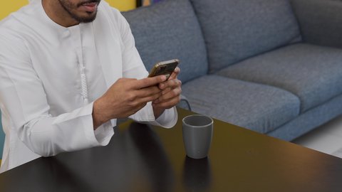 الاستمتاع بالمشروبات الساخنه ، قضاء وقت ممتع ، الدردشة عبر الانترنت ،دمج التكنولوجيا الحديثة فى الحياة اليومية ، تصوير لشاب عربي خليجي إماراتي يرتدي الكندورة والغترة البيضاء يجلس على اريكة رمادية ، يستخدم جهاز الهاتف المحمول، الاطمئنان على الاهل والاصدقاء