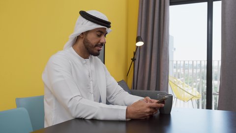 الدردشة عبر الانترنت ،دمج التكنولوجيا الحديثة فى الحياة اليومية ، تصوير لشاب عربي خليجي إماراتي يرتدي الكندورة والغترة البيضاء يجلس على اريكة رمادية ، يستخدم جهاز الهاتف المحمول، الاطمئنان على الاهل والاصدقاء ، الاستمتاع بالمشروبات الساخنه ، قضاء وقت ممتع