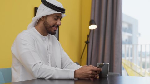 دمج التكنولوجيا الحديثة فى الحياة اليومية ، تصوير لشاب عربي خليجي إماراتي يرتدي الكندورة والغترة البيضاء يجلس على اريكة رمادية ، يستخدم جهاز الهاتف المحمول، الاطمئنان على الاهل والاصدقاء ،الدردشة عبر الانترنت ، الاستمتاع بالمشروبات الساخنه ، قضاء وقت ممتع