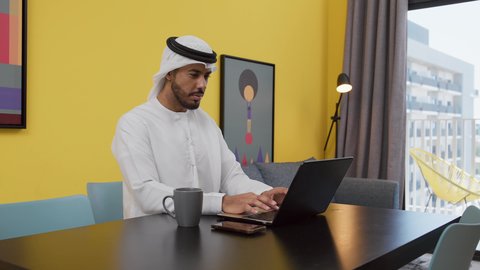 Having a good time, completing projects and tasks at home, a picture of a young Emirati Gulf Arab man wearing a white kandura and ghutra sitting on a gray sofa, using a laptop, enjoying hot drinks, the concept of technology and the use of modern technical devices