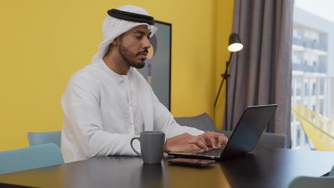 انجاز المشاريع والمهام في المنزل ،تصوير لشاب عربي خليجي إماراتي يرتدي الكندورة والغترة البيضاء يجلس على اريكة رمادية ، يستخدم جهاز الحاسوب المحمول ، قضاء وقت ممتع ,الاستمتاع بالمشروبات الساخنه ، مفهوم التكنولوجيا وإستخدام أجهزة تقنية حديثة