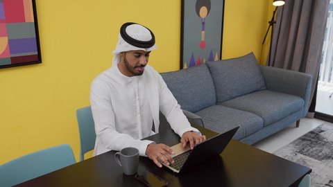 A photo of a young Emirati Gulf Arab man wearing a white kandura and ghutra sitting on a gray sofa, using a laptop, having a good time, enjoying hot drinks, completing projects and tasks at home, the concept of technology and the use of modern technical devices