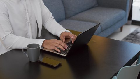 قضاء وقت ممتع ,الاستمتاع بالمشروبات الساخنه ، انجاز المشاريع والمهام في المنزل ، تصوير لشاب عربي خليجي إماراتي يرتدي الكندورة والغترة البيضاء يجلس على اريكة رمادية ، يستخدم جهاز الحاسوب المحمول ، مفهوم التكنولوجيا وإستخدام أجهزة تقنية حديثة