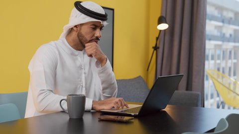 تعابير الاندماج والتركيز، انجاز المشاريع والمهام في المنزل ، تصوير لشاب عربي خليجي إماراتي يرتدي الكندورة والغترة البيضاء يجلس على اريكة رمادية ، يستخدم جهاز الحاسوب المحمول ، الاستمتاع بالمشروبات الساخنه ، قضاء وقت ممتع , مفهوم التكنولوجيا وإستخدام أجهزة تقنية حديثة