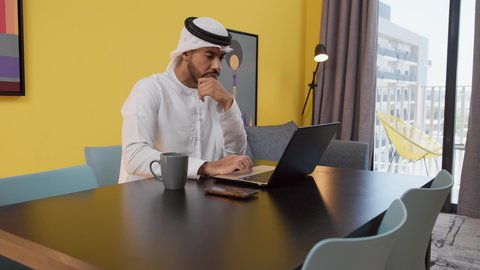 انجاز المشاريع والمهام في المنزل ، تصوير لشاب عربي خليجي إماراتي يرتدي الكندورة والغترة البيضاء يجلس على اريكة رمادية ، يستخدم جهاز الحاسوب المحمول ، الاستمتاع بالمشروبات الساخنه ، قضاء وقت ممتع , مفهوم التكنولوجيا وإستخدام أجهزة تقنية حديثة