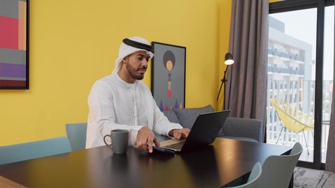 تصوير لشاب عربي خليجي إماراتي يرتدي الكندورة والغترة البيضاء يجلس على اريكة رمادية ، يستخدم جهاز الحاسوب المحمول ، الاستمتاع بالمشروبات الساخنه ، قضاء وقت ممتع , مفهوم التكنولوجيا وإستخدام أجهزة تقنية حديثة ، انجاز المشاريع والمهام في المنزل