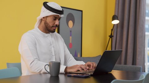 الاستمتاع بالمشروبات الساخنه ، قضاء وقت ممتع , مفهوم التكنولوجيا وإستخدام أجهزة تقنية حديثة ، انجاز المشاريع والمهام في المنزل ، تصوير لشاب عربي خليجي إماراتي يرتدي الكندورة والغترة البيضاء يجلس على اريكة رمادية ، يستخدم جهاز الحاسوب المحمول