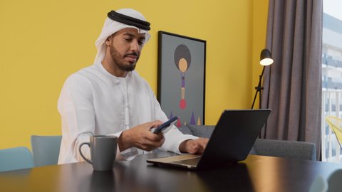 قضاء وقت ممتع , مفهوم التكنولوجيا وإستخدام أجهزة تقنية حديثة ، انجاز المشاريع والمهام في المنزل ، تصوير لشاب عربي خليجي إماراتي يرتدي الكندورة والغترة البيضاء يجلس على اريكة رمادية ، يستخدم جهاز الحاسوب المحمول ، الاستمتاع بالمشروبات الساخنه