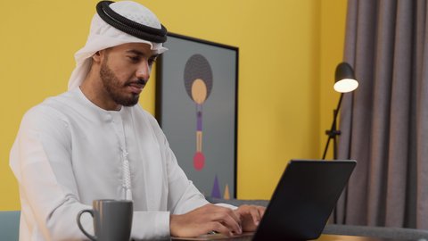 مفهوم التكنولوجيا وإستخدام أجهزة تقنية حديثة ، انجاز المشاريع والمهام في المنزل ، قضاء وقت ممتع ,تصوير لشاب عربي خليجي إماراتي يرتدي الكندورة والغترة البيضاء يجلس على اريكة رمادية ، يستخدم جهاز الحاسوب المحمول ، الاستمتاع بالمشروبات الساخنه