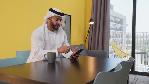 تصوير لشاب عربي خليجي إماراتي يرتدي الكندورة والغترة البيضاء يجلس على اريكة رمادية ، يستخدم جهاز الهاتف المحمول، دمج التكنولوجيا الحديثة فى الحياة اليومية ، الاطمئنان على الاهل والاصدقاء ،الدردشة عبر الانترنت ، الاستمتاع بالمشروبات الساخنه ، قضاء وقت ممتع