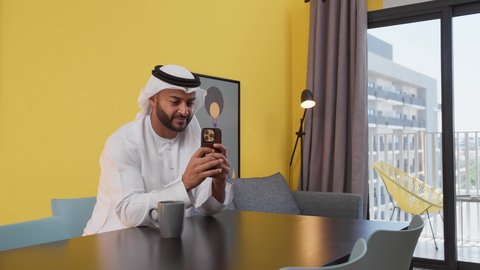 توثيق اللحظات التذكارية، دمج التكنولوجيا الحديثة فى الحياة اليومية ، الاستمتاع بالمشروبات الساخنه ، قضاء وقت ممتع ، تصوير لشاب عربي خليجي إماراتي يرتدي الكندورة والغترة البيضاء يجلس على اريكة رمادية ، يستخدم جهاز الهاتف المحمول