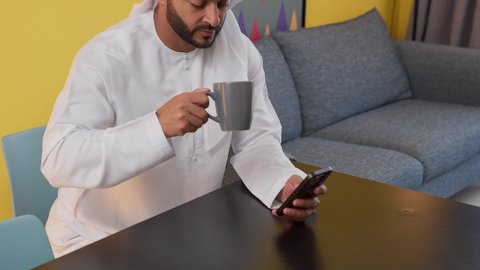 الاطمئنان على الاهل والاصدقاء ،الدردشة عبر الانترنت ، الاستمتاع بالمشروبات الساخنه ، قضاء وقت ممتع ، تصوير لشاب عربي خليجي إماراتي يرتدي الكندورة والغترة البيضاء يجلس على اريكة رمادية ، يستخدم جهاز الهاتف المحمول، مفهوم التكنولوجيا وإستخدام أجهزة تقنية حديثة