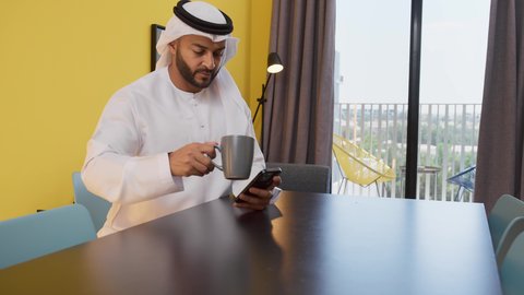 الاستمتاع بالمشروبات الساخنه ، قضاء وقت ممتع ، تصوير لشاب عربي خليجي إماراتي يرتدي الكندورة والغترة البيضاء يجلس على اريكة رمادية ، يستخدم جهاز الهاتف المحمول، مفهوم التكنولوجيا وإستخدام أجهزة تقنية حديثة ، الاطمئنان على الاهل والاصدقاء ،الدردشة عبر الانترنت