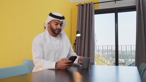 A photo of a young Emirati Gulf Arab man wearing a white kandura and ghutra sitting on a gray sofa, using a mobile phone, the concept of technology and the use of modern technical devices, checking on family and friends, chatting online, enjoying hot drinks, and having a good time.