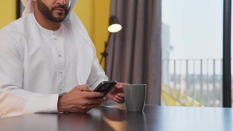 مفهوم التكنولوجيا وإستخدام أجهزة تقنية حديثة ، الاطمئنان على الاهل والاصدقاء ،الدردشة عبر الانترنت ، الاستمتاع بالمشروبات الساخنه ، قضاء وقت ممتع ، تصوير لشاب عربي خليجي إماراتي يرتدي الكندورة والغترة البيضاء يجلس على اريكة رمادية ، يستخدم جهاز الهاتف المحمول