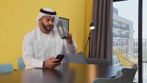 الدردشة عبر الانترنت ، الاستمتاع بالمشروبات الساخنه ، قضاء وقت ممتع ، مفهوم التكنولوجيا وإستخدام أجهزة تقنية حديثة ، الاطمئنان على الاهل والاصدقاء ، تصوير لشاب عربي خليجي إماراتي يرتدي الكندورة والغترة البيضاء يجلس على اريكة رمادية ، يستخدم جهاز الهاتف المحمول