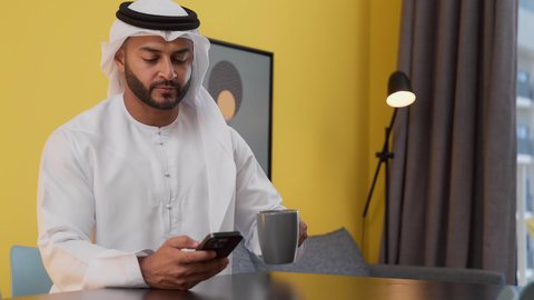 قضاء وقت ممتع ، مفهوم التكنولوجيا وإستخدام أجهزة تقنية حديثة ، الاطمئنان على الاهل والاصدقاء ، الدردشة عبر الانترنت ، الاستمتاع بالمشروبات الساخنه ، تصوير لشاب عربي خليجي إماراتي يرتدي الكندورة والغترة البيضاء يجلس على اريكة رمادية ، يستخدم جهاز الهاتف المحمول